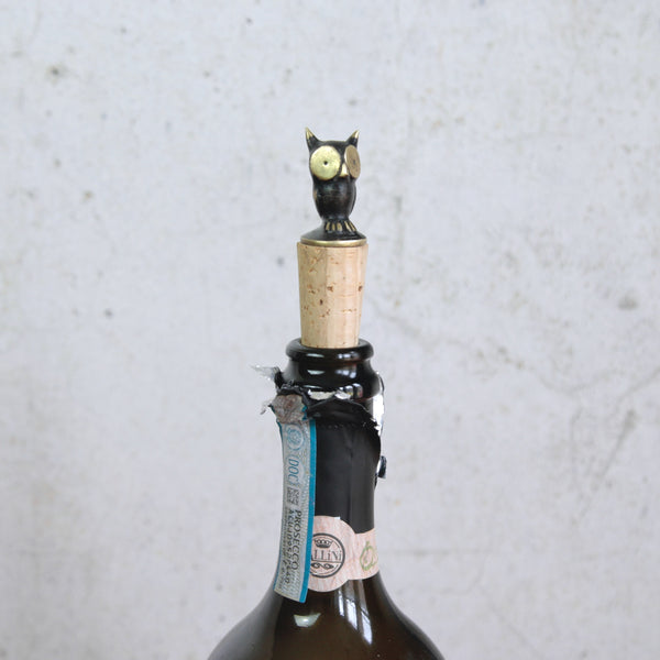 Wine bottle with an owl-shaped bottle stopper on a textured gray background. The Mid-century brass owl bottle stopper is designed by Walter Bosse for Herta Baller, Vienna. A sought-after collectible blending art with function.
