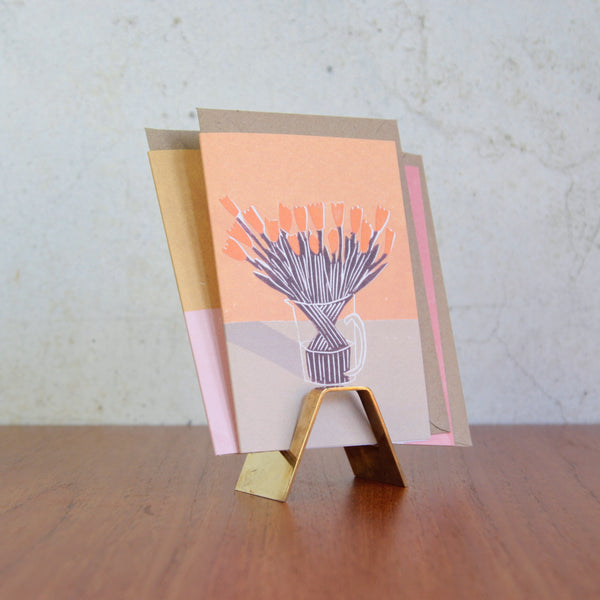 Artistic mood image of a Modernist brass card holder by Karl Hagenauer, sleek Viennese design circa 1950. The card holder contains a greeting card with a floral design.