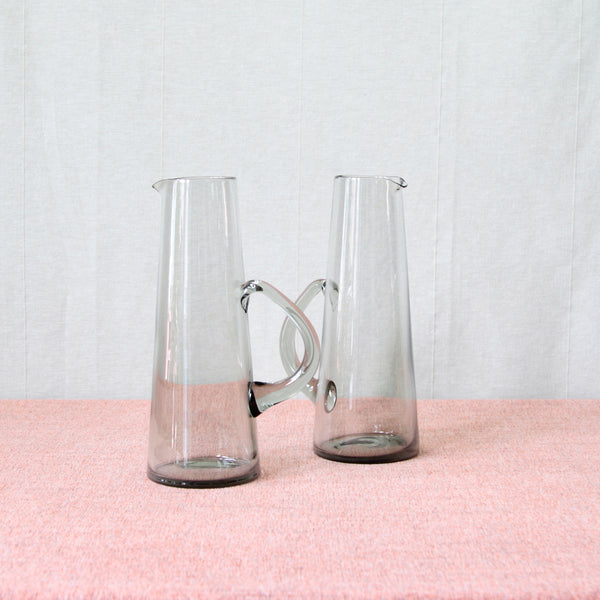 A pair of cocktail pitchers with conical bodies, hand-pulled pouring spouts, and biomorphic handles. Designed in 1957 by Per Lütken as part of his series of 3 'Martini' pitchers. These examples, with a height of 24.5 cm, are both medium version from the series. The greenish-grey colour of the glass is known as 'Smoke', as was Lütken's signature colour way.