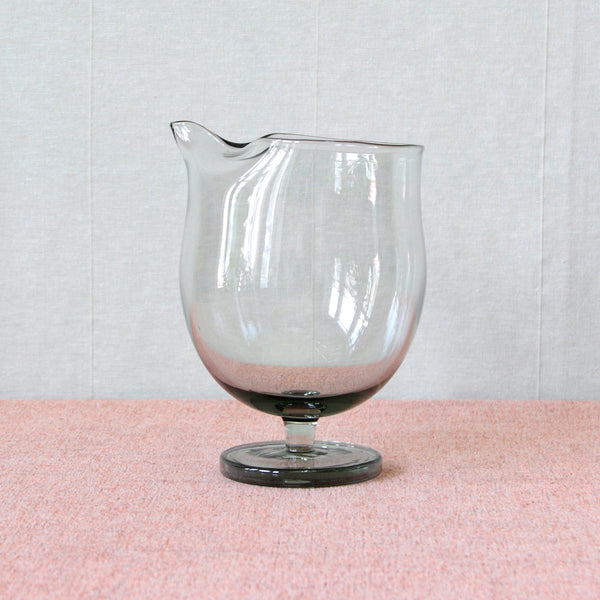 A footed bulbous bodied jug for mixing cocktails. This fun ball shaped design by Per Lütken from 1958 was inspired by Tivoli Gardens amusement park. The greenish-grey colour of the glass is known as 'Smoke', and was Lütken's signature colour way at Holmegaard.