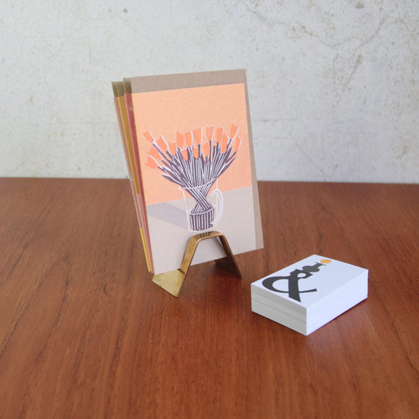 Top down artistic mood shot showing a collectible Hagenauer Vienna brass card stand. A card with a floral design is positioned in the stand whilst to the right is a pile of Art & Utility business cards which could also be displayed in the Hagenauer rack.