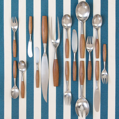 An artistic image showing, from the top down, sixteen pieces of 'Fjord' cutlery designed by Jens Quistgaard for Dansk Designs. The handles of the cutlery are solid Siamese teak, the blades are high quality stainless steel.