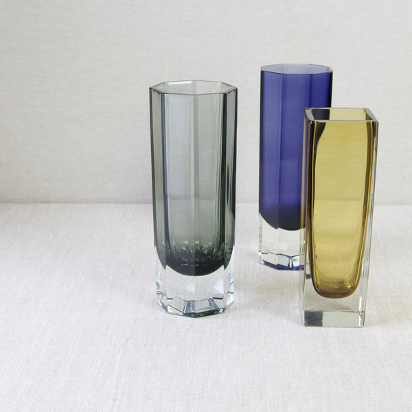 Top down artistic mood shot showing three Nuutajarvi Notsjo glass vases with geometric forms stood grouped together. Each is a different colour, namely grey, purple, and yellow. However, all 3 were designed by Kaj Franck one of the leading figures in 20th century Scandinavian design history.