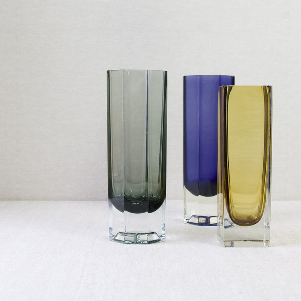 Head on low down mood shot showing 3 Kaj Franck vases all with clean and sharp geometric designs stood together. Left to right the colours are grey, purple and yellow.