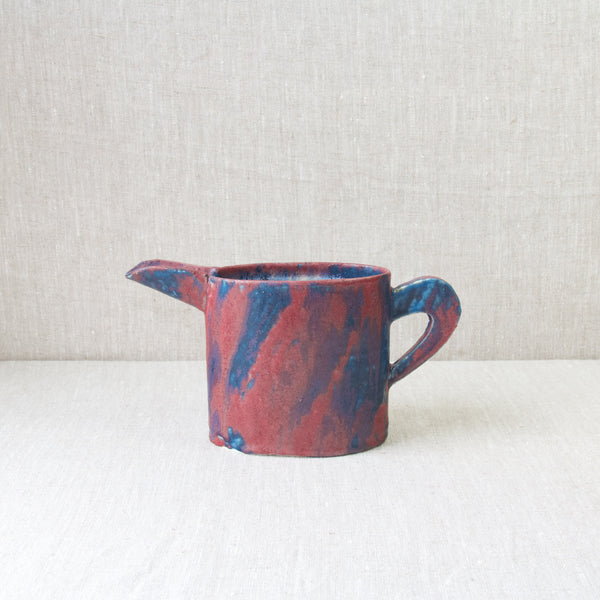 An Emmanuel Cooper jug with an experimental red and blue glaze with splashes of purple. The jug not unlike forms explored by Walter Keeler stands atop a plain linen tablecloth.