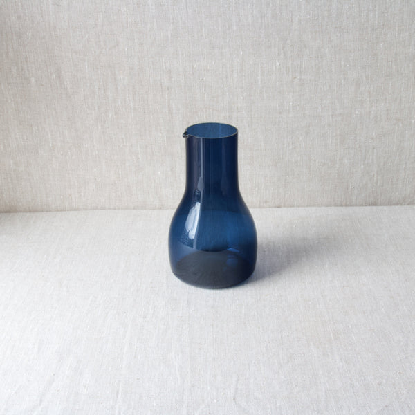 Top down view of a deep indigo blue glass model 1615 pitcher designed by Kaj Franck. This rare piece of Scandinavian design, that was produced for just one year in 1963, stands atop a linen tablecloth and the backdrop is also a neutral cloth.