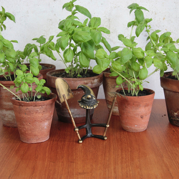 Fun and artistic mood shot showing a collectible mid-century Walter Bosse seated gnome ornament with gardening tools stood between potted basil plants.