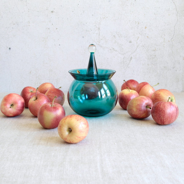 Artistic still life of glass with fruit. A Nanny Still Harlekiini glass bowl with lid, produced by Riihimäen Lasi, featuring bold turquoise colour and geometric form.