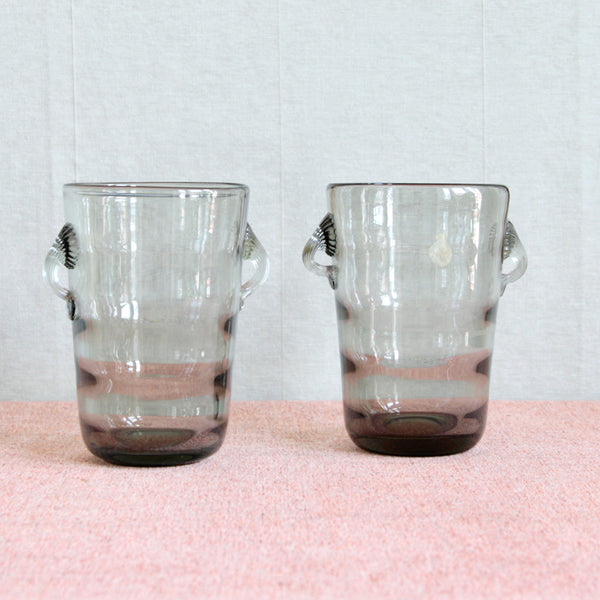 Two Danish glass ice buckets with handles on a pink surface and light grey background. The Modernist designs are optic-moulded glass. Design by Jacob Bang, 1937, and 'Smoke' grey colour by Per Lütken, 1954. Produced by Holmegaard.
