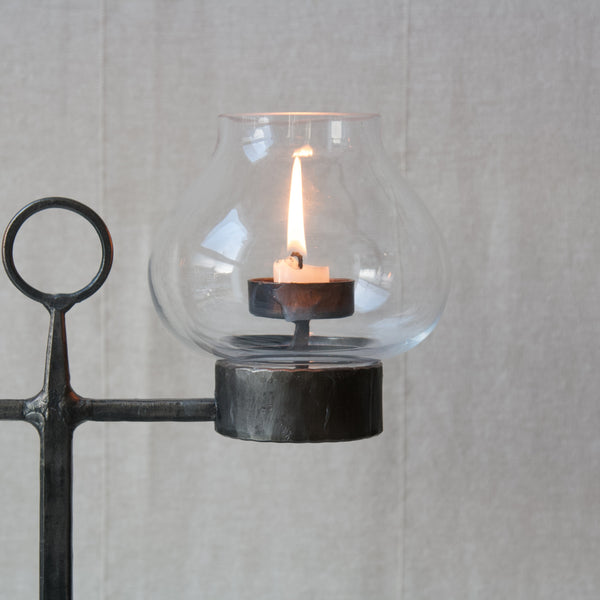 Zoomed in image showing a canlde behind a clear glass hurricane lampshade shaped like a tulip flower. This design is by Bertil Vallien. The candelabrum is stable enough for outdoor use.