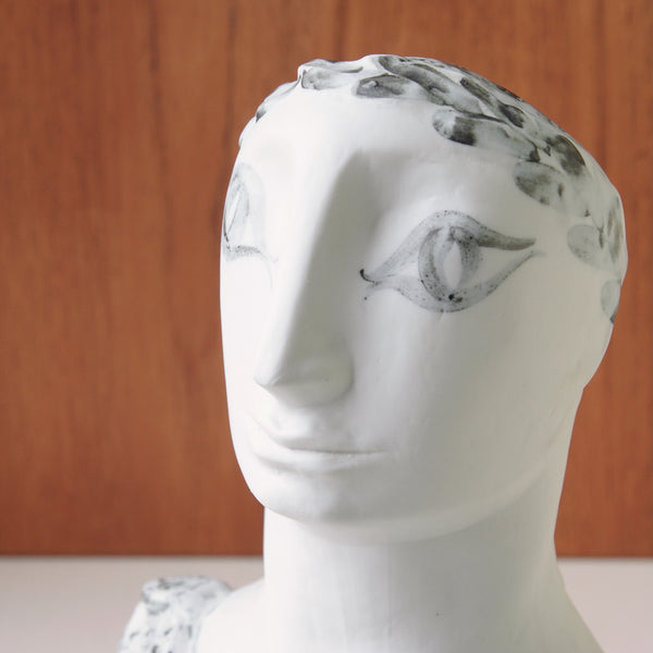 White marble like bust of a Grecian man against a wooden panel background. Vintage Susan Parkinson sculpture capturing the noble serenity of a Greek figure, a hallmark of modern British ceramic design.