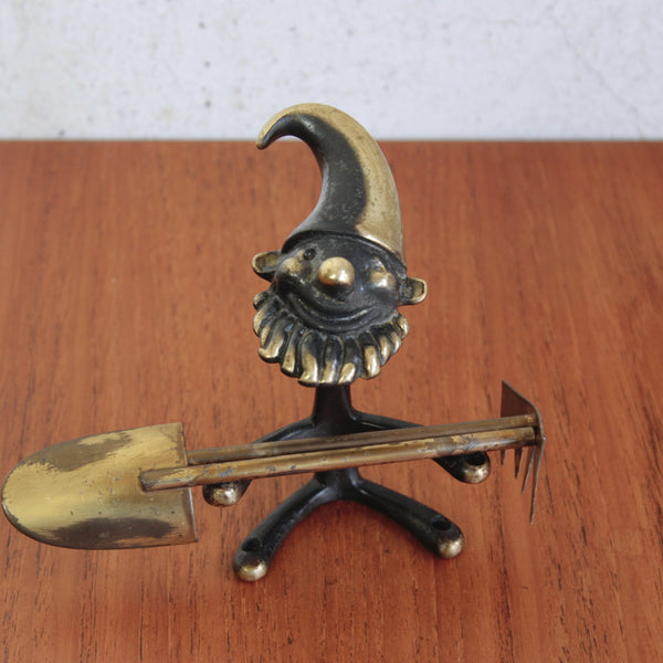 Decorative metal figure with a hat and tools on a wooden surface. This Walter Bosse vintage gnome with rake and spade is a charming collectible for gardeners and design lovers.