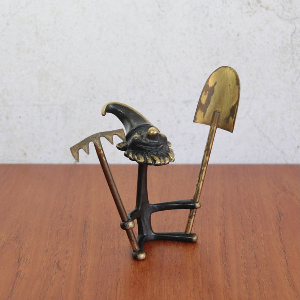 Decorative figurine of a gnome with a shovel and rake on a wooden surface. Walter Bosse gardening gnome ornament, holding rake and spade, mid-century brass collectible, circa 1965.