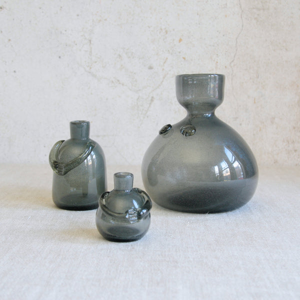 Artistic mood shot showing three iridescent carborundum glass vases of different sizes on a textured linen surface. Rare early Erik Höglund vases from Boda, Sweden. Named people decanters / vases / candlesticks each of designs displays a bold figurative silhouette. Rare Nordic collectible.