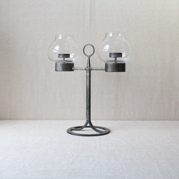 Artful image showing a tall black iron candelabrum with two glass shades stood atop a pale beige linen table cloth. The design is by Bertil Vallien for Boda Smide.