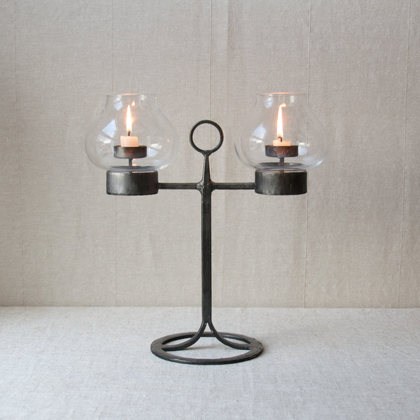 A straight on mood image showing a Bertil Vallien candelabrum complete with two lit dinner candles. The candles sit behind clear glass hurricane shades which shelter them from the wind. This means this item available from Art & Utility London, could be used outside on the garden table on the patio.