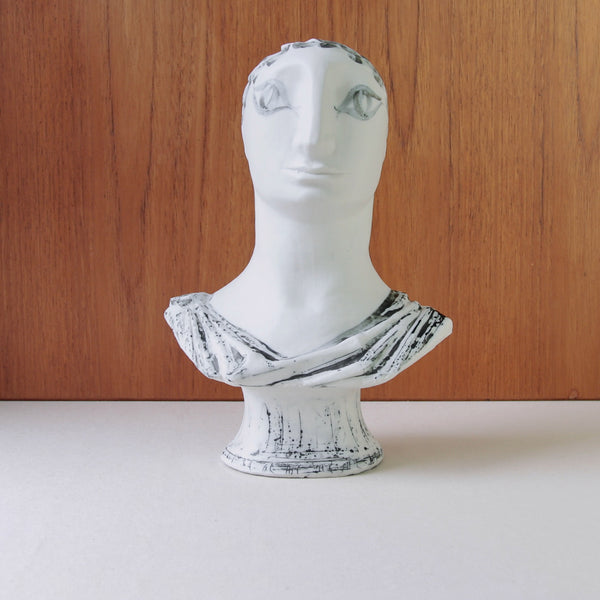 White ceramic bust with face on a wooden background. 1950s slip-cast porcelain sculpture by Susan Parkinson with laurel wreath and column base, inspired by Picasso’s neoclassicism.
