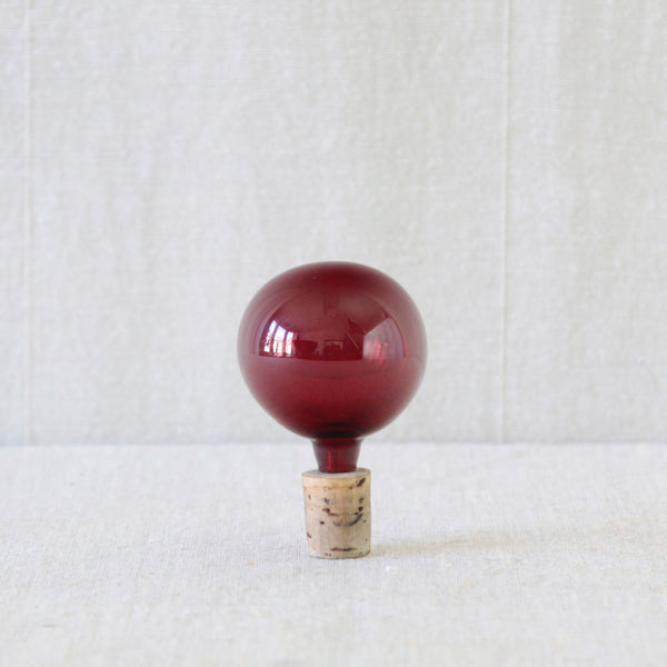 A red glass decanter stopper with a rounded bulbous top and a natural cork base, standing upright. This brilliant ruby-red spherical stopper crowns a Kaj Franck Kremlin Bells / Kellot decanter set.