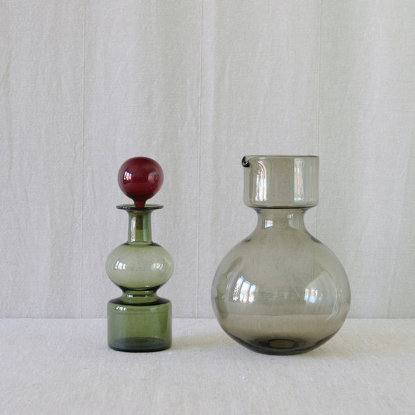 Kremlin Kellot by Kaj Franck, seperated into two sections. The pitcher is in smoke-grey, whereas the decanter is in bottle-green with a ruby-red glass ball stopper. A most collectible example of Finnish Modernist design.