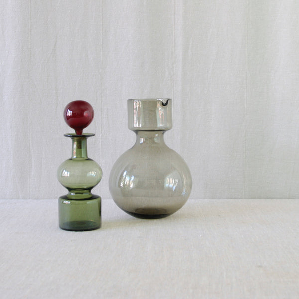 Two vintage glass vessels that form a sculptural Kremlin Kellot decanter and pitcher by Kaj Franck. The decanter is in green with a dark red spherical stopper, and the other is a large, bulb-shaped grey glass pitcher. The contrasting colours by Nuutajärvi Notsjö emphasis the designs distinctive form when stacked.