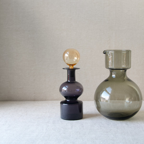 Top half of a 'Kremlin Kellot' in purple and yellow, stood to the left of the bottom half of a 'Kremlin Bells' carafe in grey. Design by leading Scandinavian designer Kaj Franck for Nuutajärvi Notsjö, the maker of the most collected glassware from Finland.