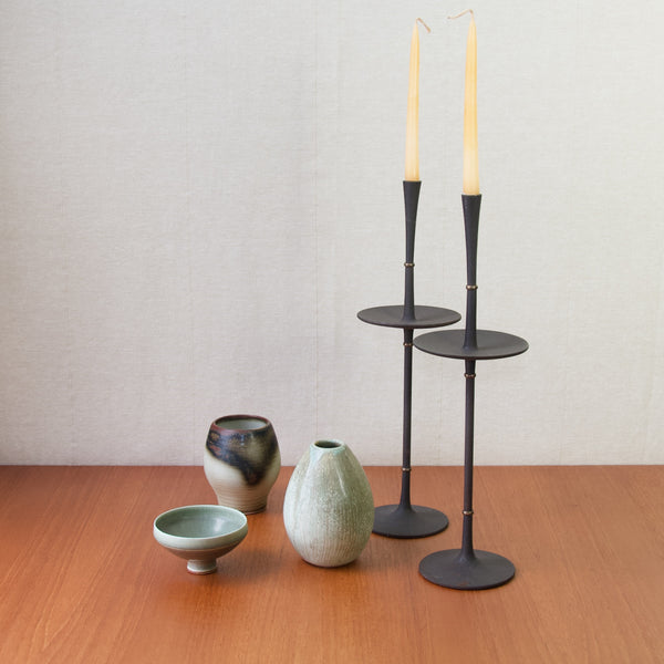 Still life mood image showing 3 ceramic vases stood to the left of a pair of tall and impressive black cast iron candlesticks by Jens Quistgaard. The ceramic vases are by Berndt Friberg, Anja Jaatinen, and Jacob Bang.