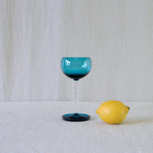 A turquoise blue and clear glass 'Harlekiini' goblet shaped wine glass  stood next to a yellow lemon for scale. Design by Nanny Still for Riihimäki.