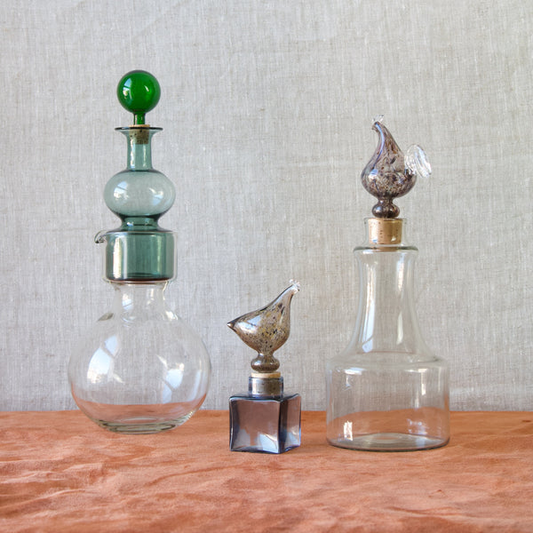 A still life mood shot showing a group of Kaj Franck decanter bottles. From left to right these are a “Kremlin Bells” Decanter and Pitcher, a “Kukkopullo” or Rooster Bottle, and a KF254 Hen Bottle. All three of these highly decorated and collected designs were produced by the expert glassmakers at Nuutajärvi Notsjö, Finland, in the mid-1950s to early-1960s. For sale in London from Art & Utility, who offer worldwide shipping.