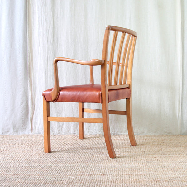 A wooden armchair with a slatted backrest, curved armrests, and a red leather seat cushion. Model 1675 design desk chair by Ole Wanscher.