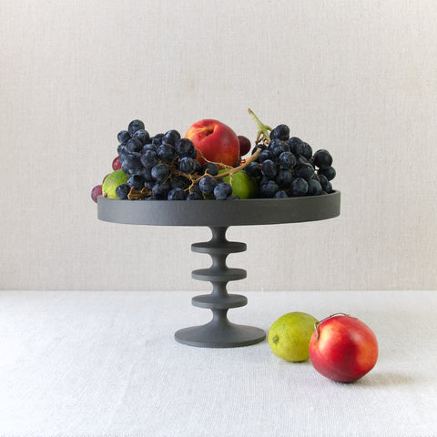 Colourful mood image showing a black cast iron Robert Welch fruit stand piled high with ripe fruit including grapes, limes, and nectarines. Classic post-war British Modernism.