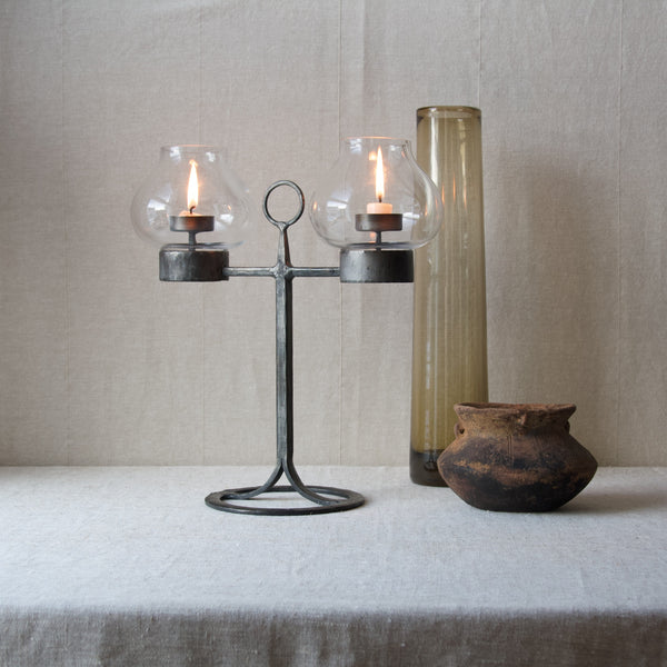 A mood image showing a mid century Swedish candelabrum by Bertil Vallien stood on a linen tablecloth next to a tall smokey brown coloured glass vase and a squat ceramic vessel of considerable age.
