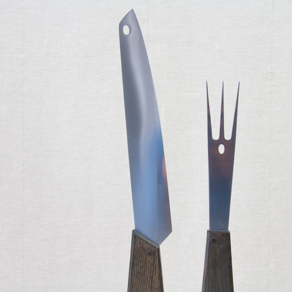 Head on shot of a carving knife and a carving fork. These are in stainless steel and belong to a set design by Tapio Wirkkala for Hackman. What makes this design special is that their handles are made from historic wood salvaged from the warship Vasa which sank in the 17th century.