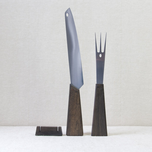 Head on shot showing a Tapio Wirkkala carving fork & knife set, with small block stand, stood up straight. This set was produced by Hackman using historic Black Oak salvaged from Vasa, a 17th century Swedish warship.