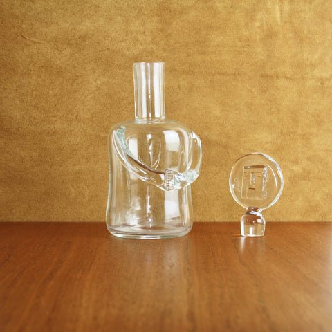 A clear Erik Hoglund scandinavian glass decanter in the shape of a person, with the head stopper removed 