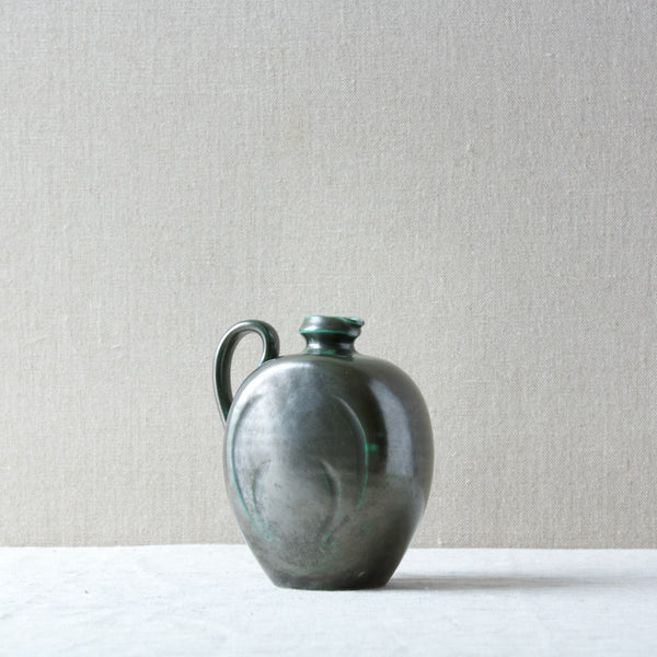 A Harald Östergren green ceramic jug standing on a natural linen table cloth in natural daylight.