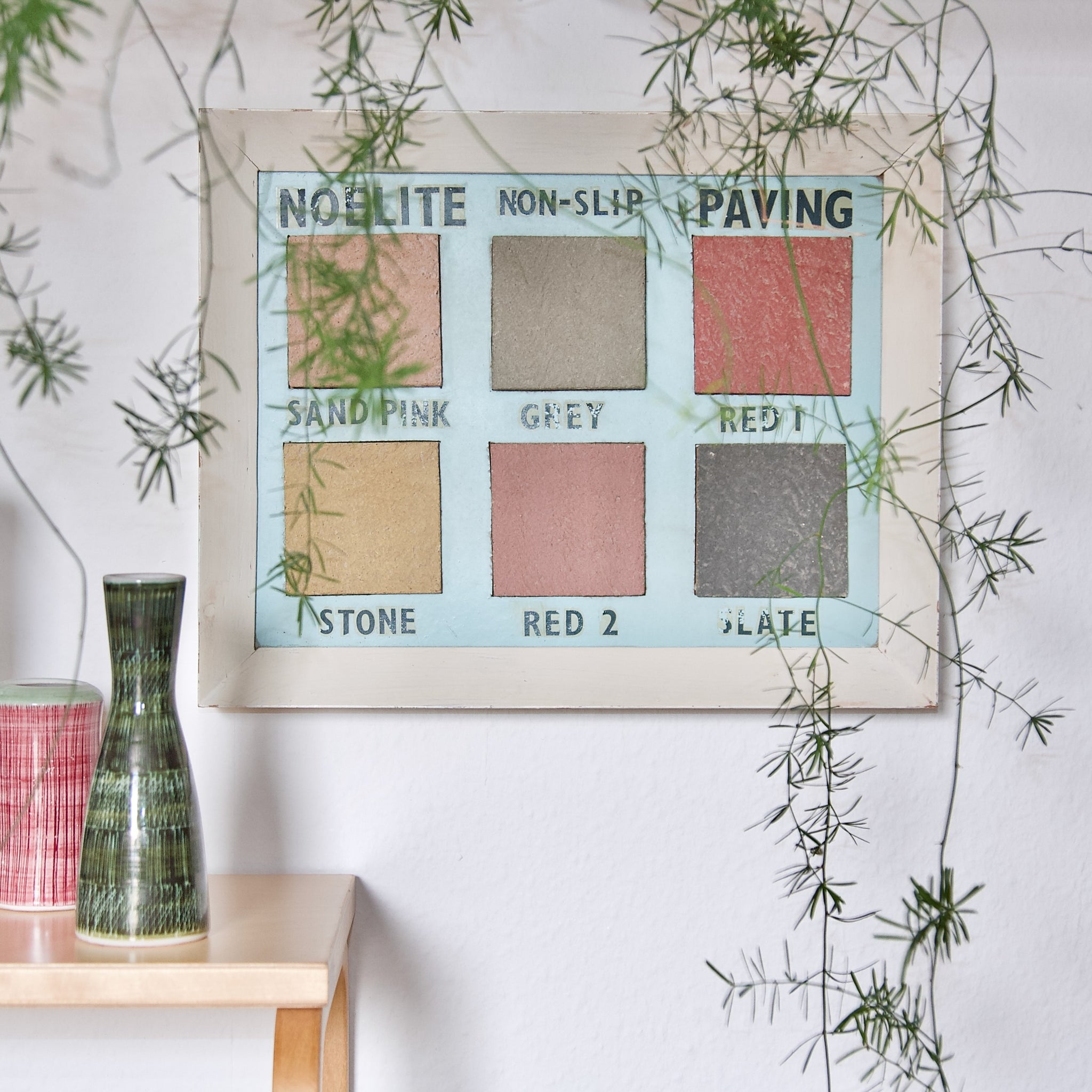 Artistic mood image showing an old vintage advertising board hung on a wall as a pice of art. Accompanying the British found art object are two sgraffito Rye pottery vases and obscuring the view is a trailing plant.