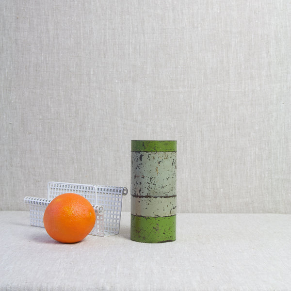 Stylised mood image showing a still life featuring two storage jars by A non-matching pair of cylindrical tins in the style of German designer and craftsperson Hans Przyrembel stood next to a white metal letter rack by Mathieu Matégot and an orange fruit. 