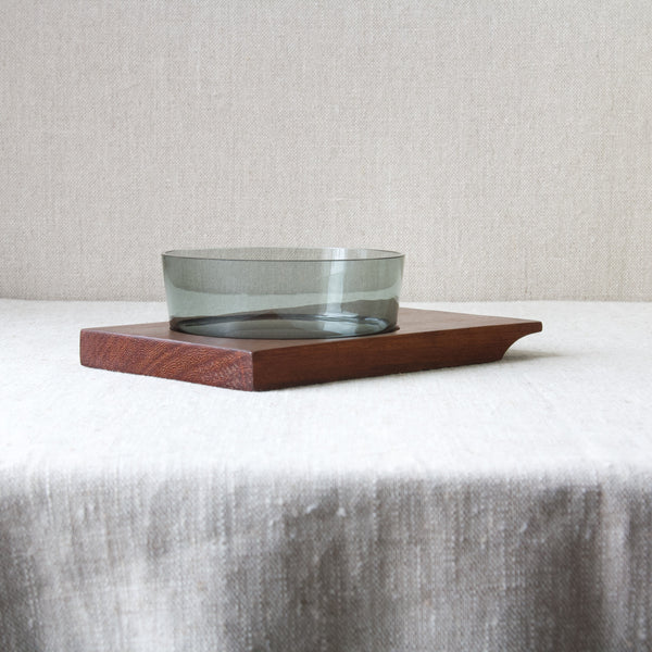 Mood image showing a smoke grey coloured glass dish stood on top of a reddish brown coloured teak wood tray. Classic Scandinavian design made by Nuutajärvi Glassworks to a design by Saara Hopea, 1955.
