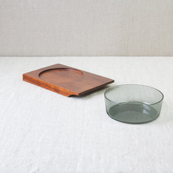 A round glass hors d'oeuvre dish stood on top of a grey linen table cloth. A rectangular teak wood tray stands close by. Designed by Saara Hopea for Nuutajärvi Glassworks, 1955.