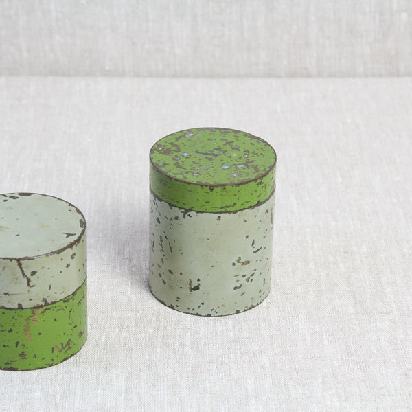 Image looking down onto the top of a cylindrical brass tin that has an enamelled exterior. The lid is vibrant green whilst the base is a putty grey. Design inspired by Hans Przyrembel.